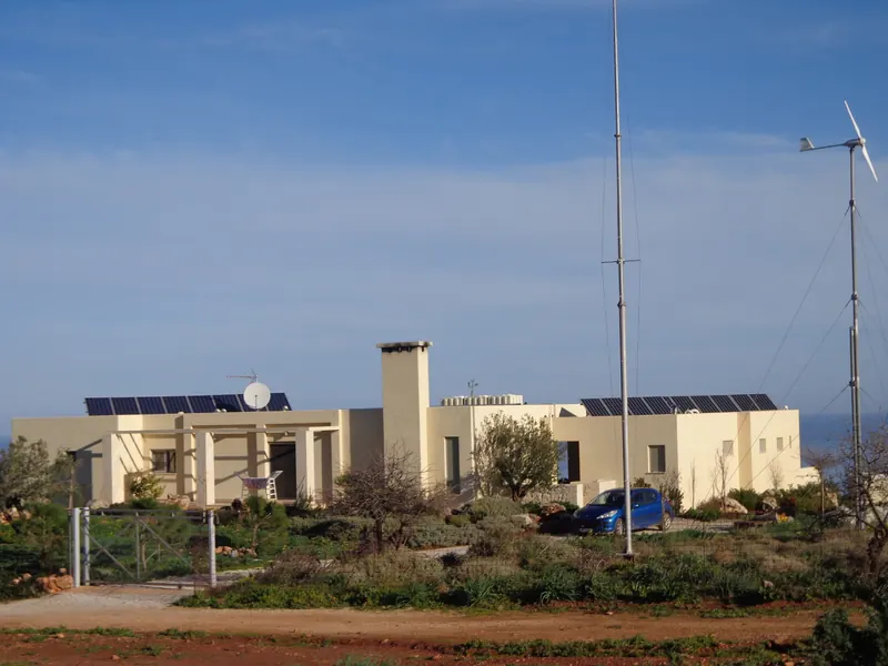 The pale yellow single-storey house with solar panels, a wind turbine and a tall mast