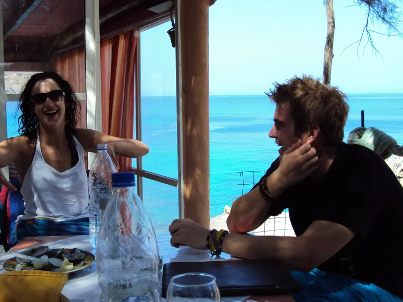 Two people laughing at a table in a seaside taverna, turquoise sea behind